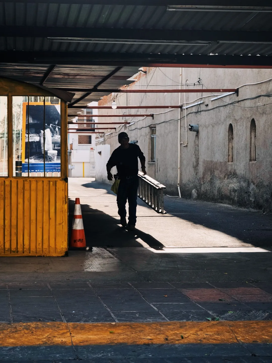 Shadowed figure walking through a covered urban passage in León, carrying a ladder through a hard patch of sunlight beside a yellow booth, concrete walls, overhead beams, and a traffic cone.